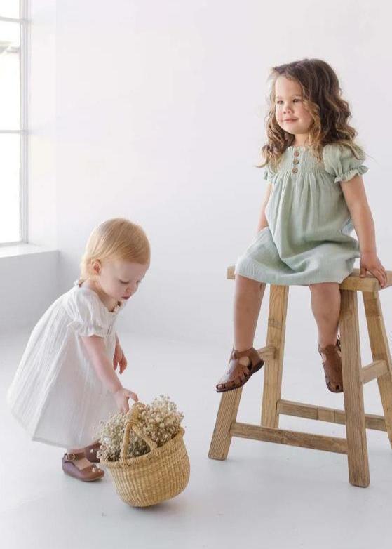 Two young children in linen dresses, one in sage green and one in white, in a bright room