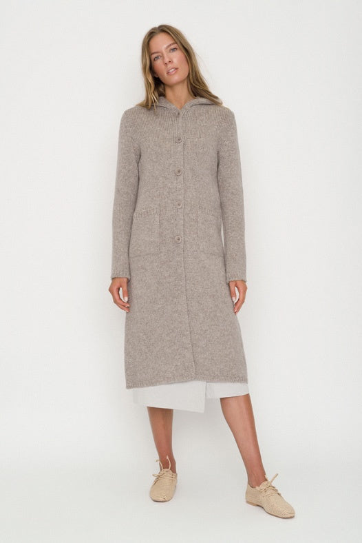 Woman wearing a long wool coat with hood and buttons down the front in natural colour.