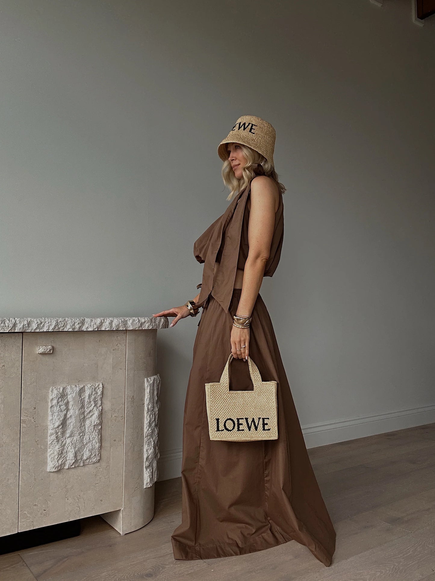 Woman in a brown sleeveless linen dress, straw hat, and straw tote bag, in a minimal room.