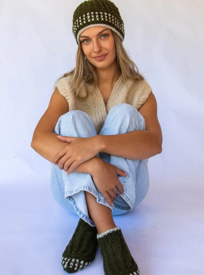 Woman wearing knitted olive booties and matching beanie, light knit vest, and jeans
