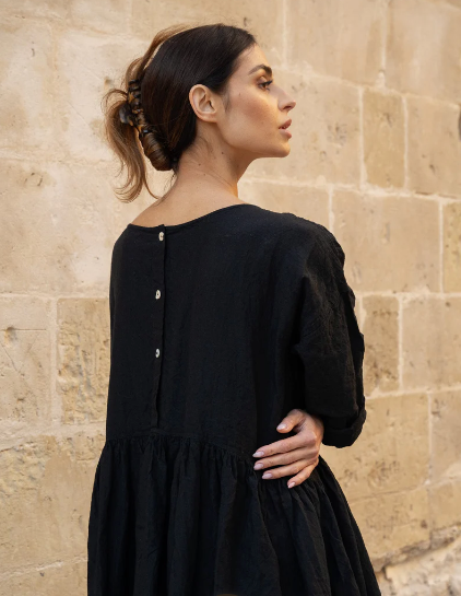 Woman wearing a black linen dress with button detail, standing against a stone wall