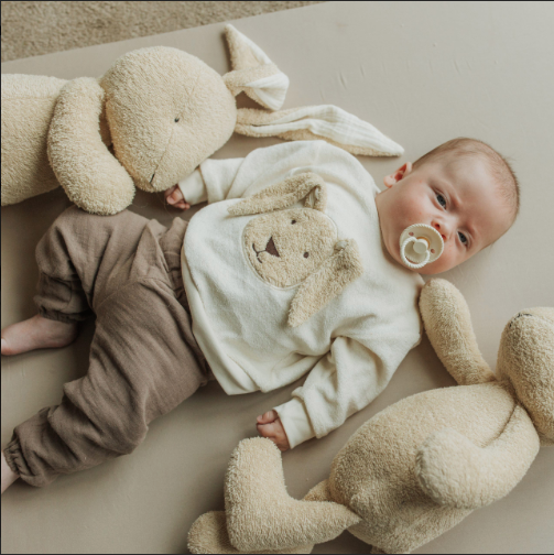 Baby wearing linen sweater and pants, surrounded by plush bunny toys, lying on a beige surface