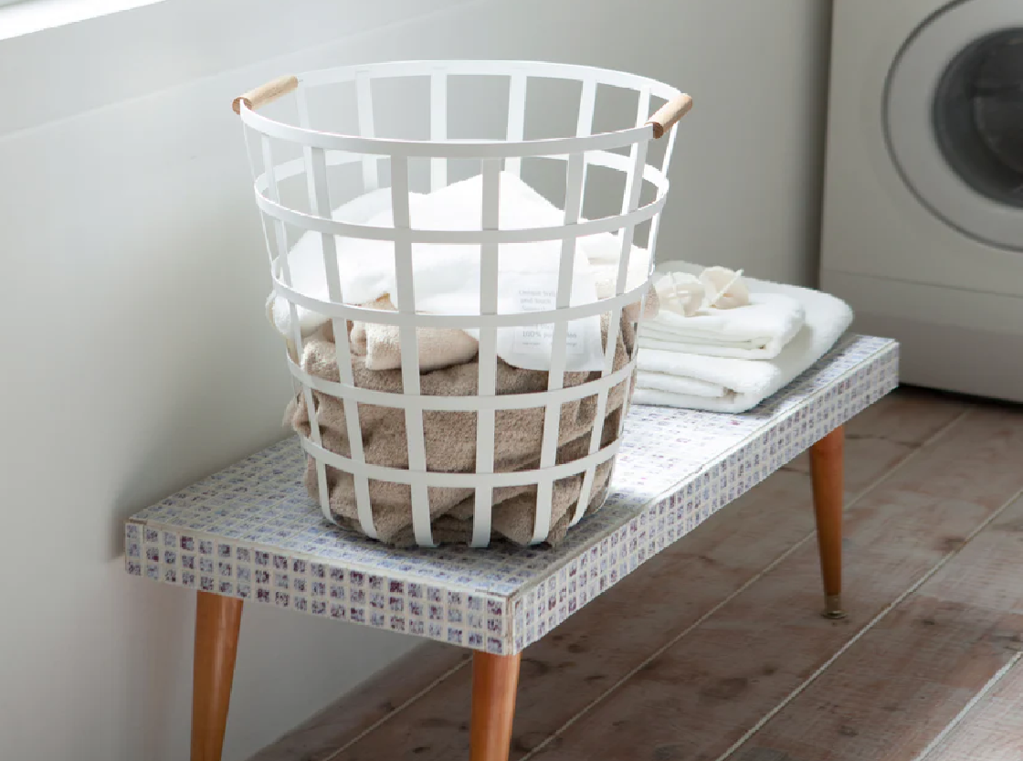White laundry basket with wooden handles filled with folded linen towels on a bench in a laundry room