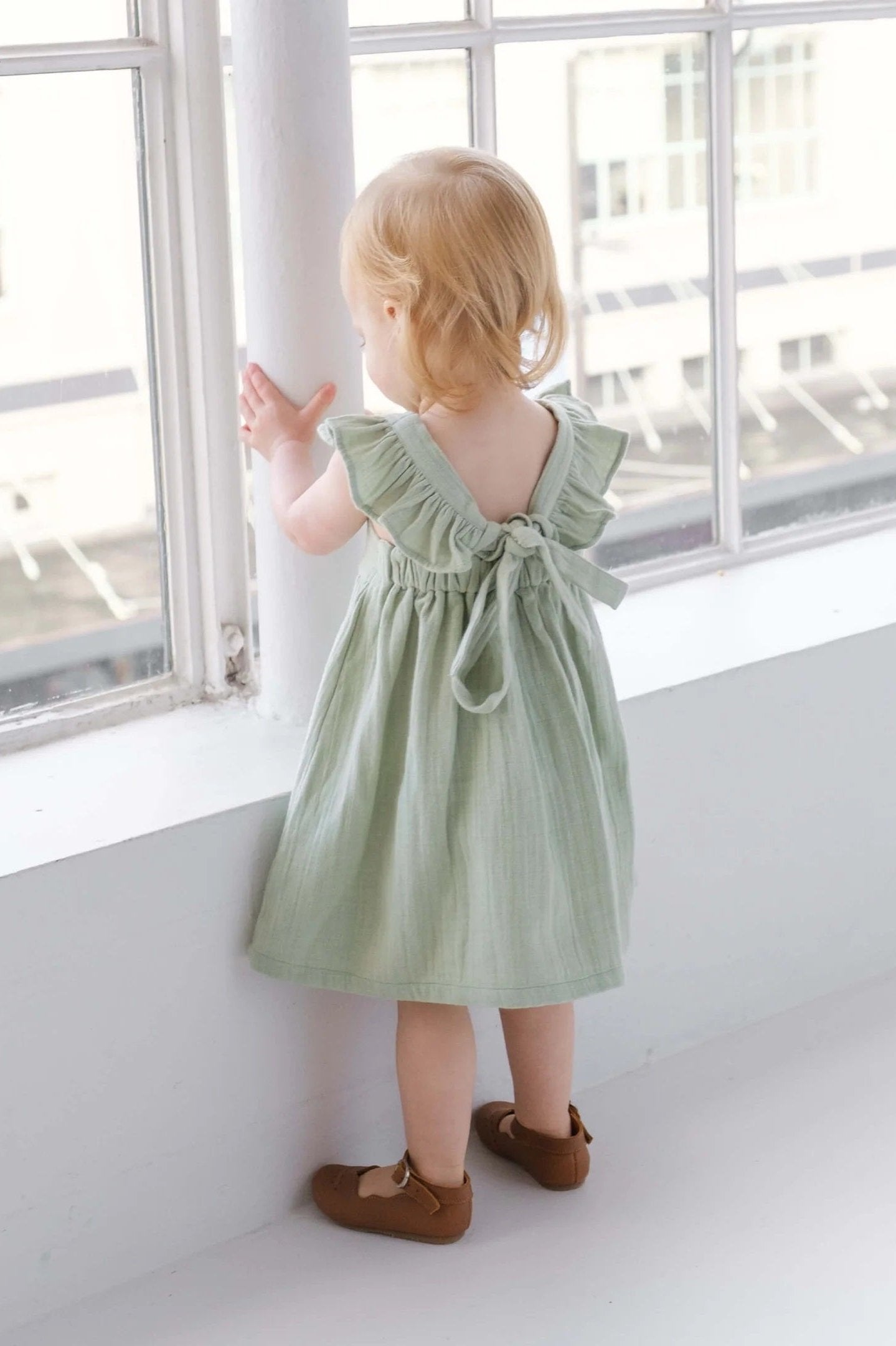 Toddler in light green linen dress with ruffle sleeves standing by bright window