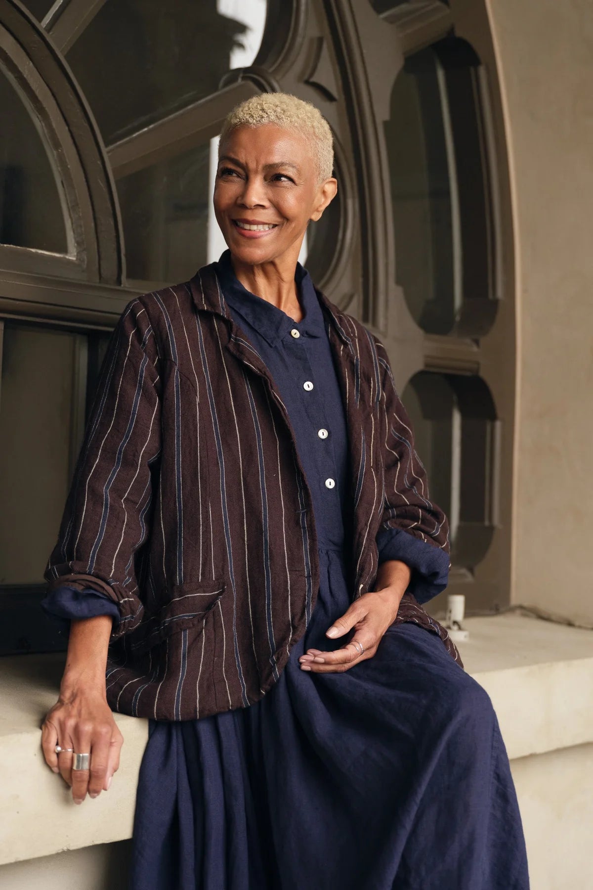 Woman wearing a brown striped linen jacket over a navy linen dress, sitting by a window.