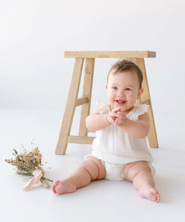 Smiling baby wearing white linen ruffle top and bloomers, sitting by wooden stool