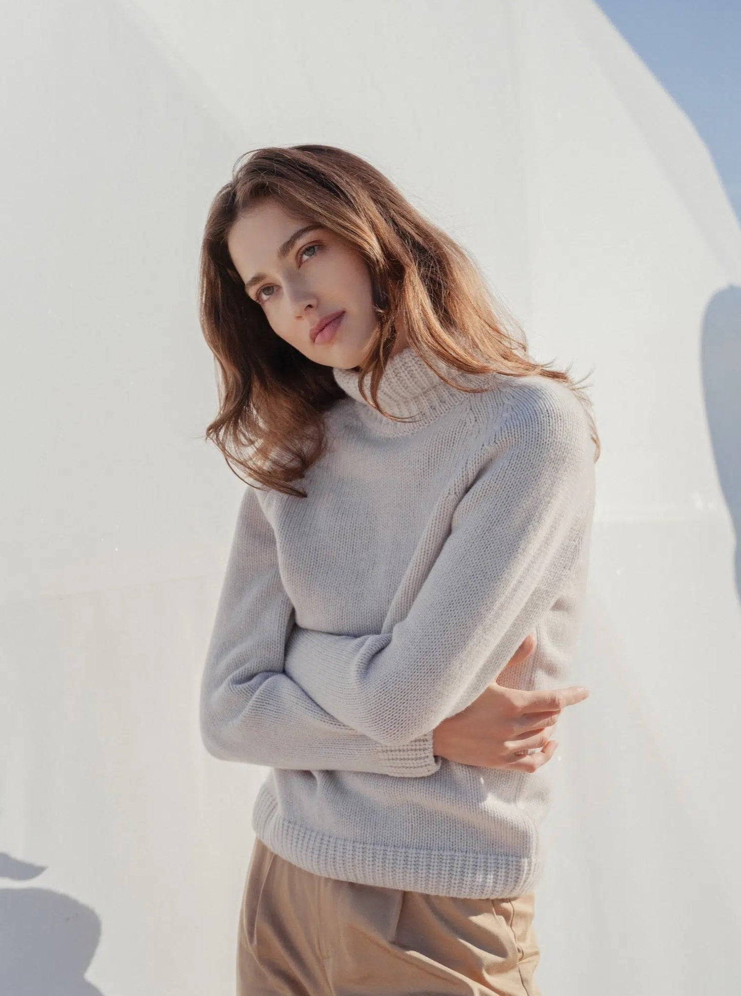 Woman in a light knit turtleneck sweater and beige pants, posing outdoors in soft sunlight