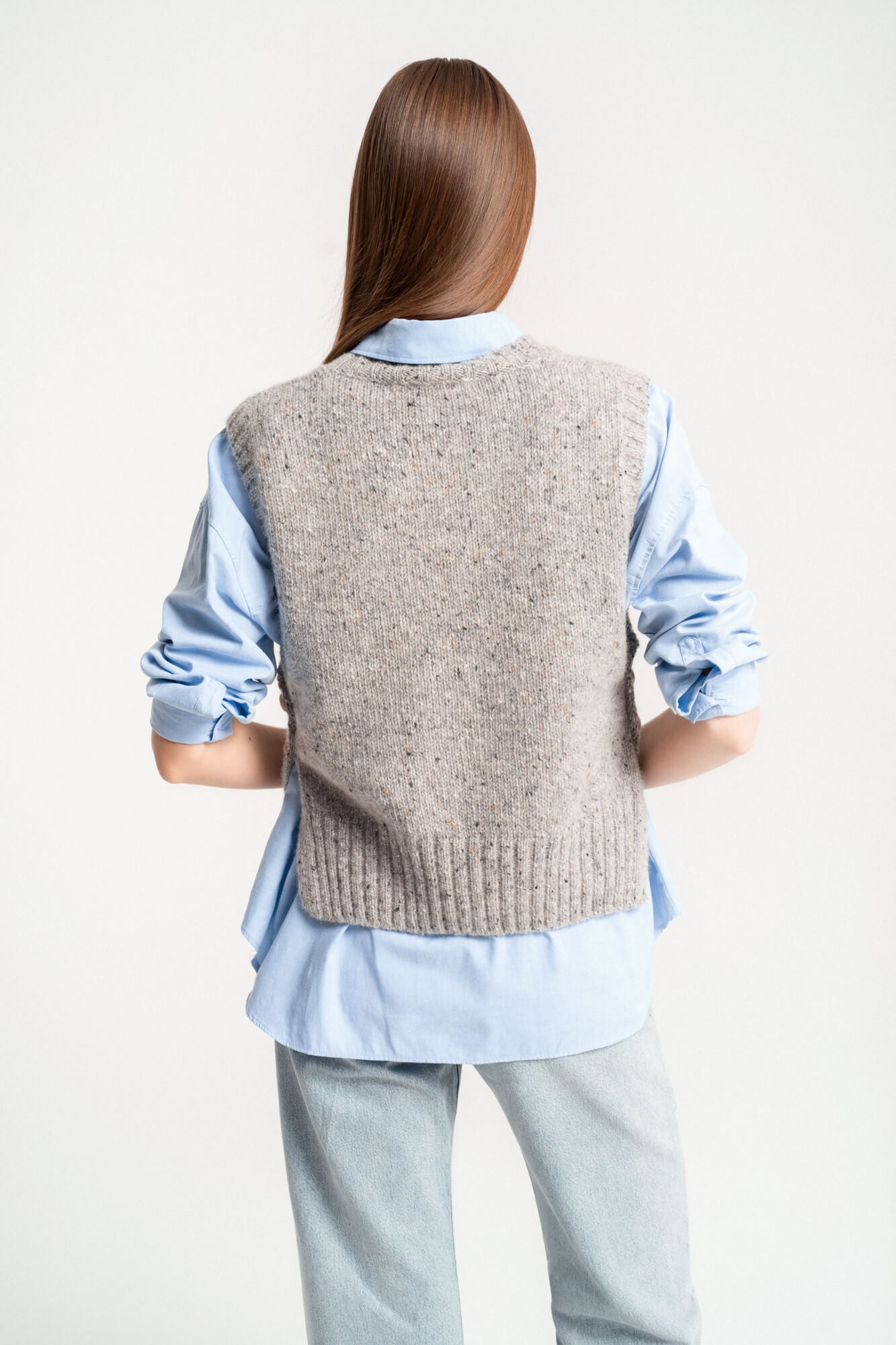 Woman wearing a grey knit vest over a light blue shirt and light denim pants, back view
