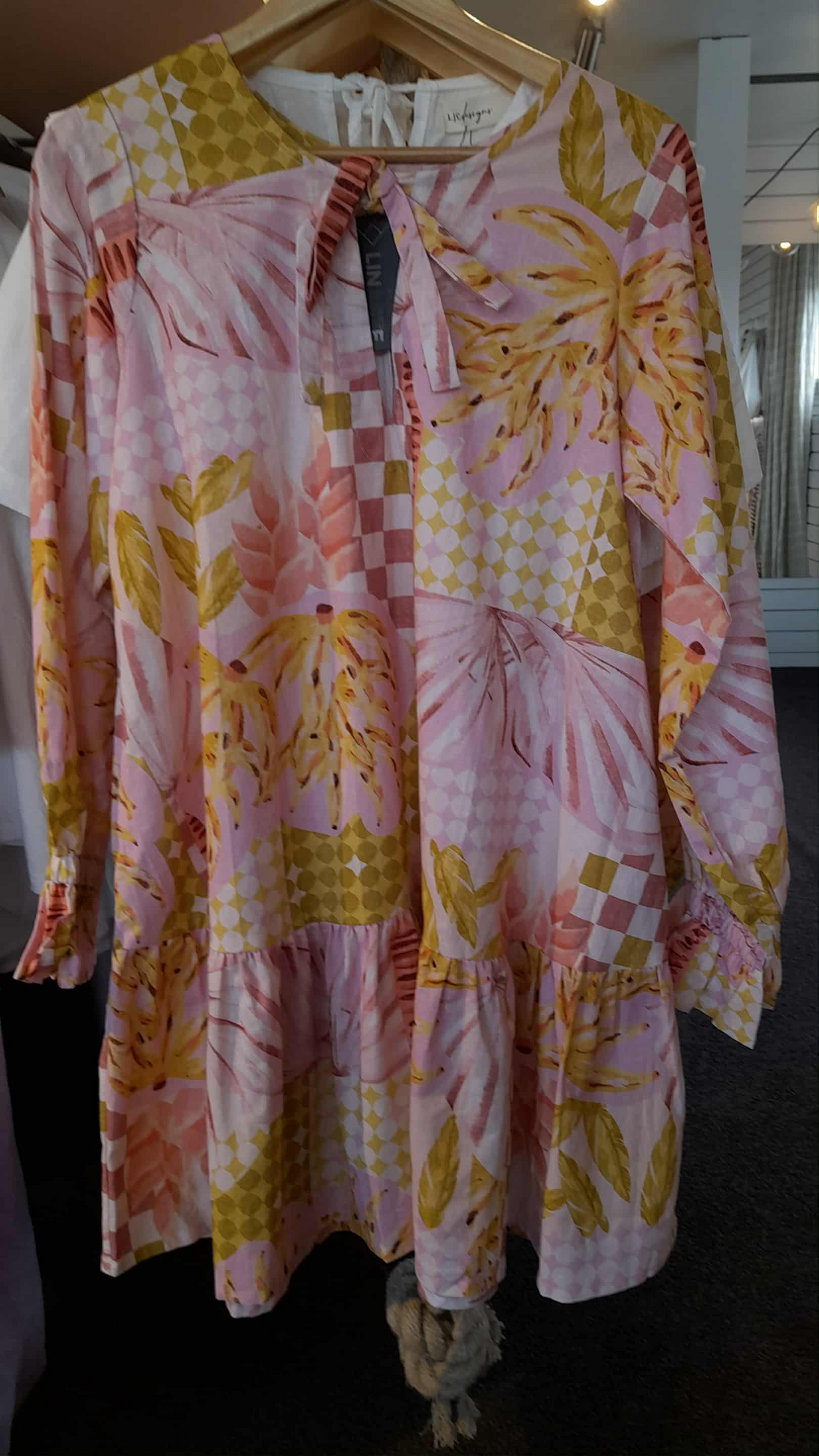 Dress on hanger is a billowy smock dress with tie neck, long sleeve with ruffle elasticated cuff in pink banana print.