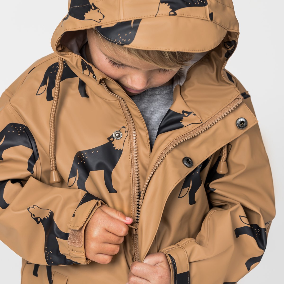 Child in tan hooded rain jacket with black wolf print, zipping up, kids outdoor clothing
