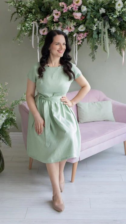 Woman in mint green linen dress with tie belt, standing by pink sofa and floral decor