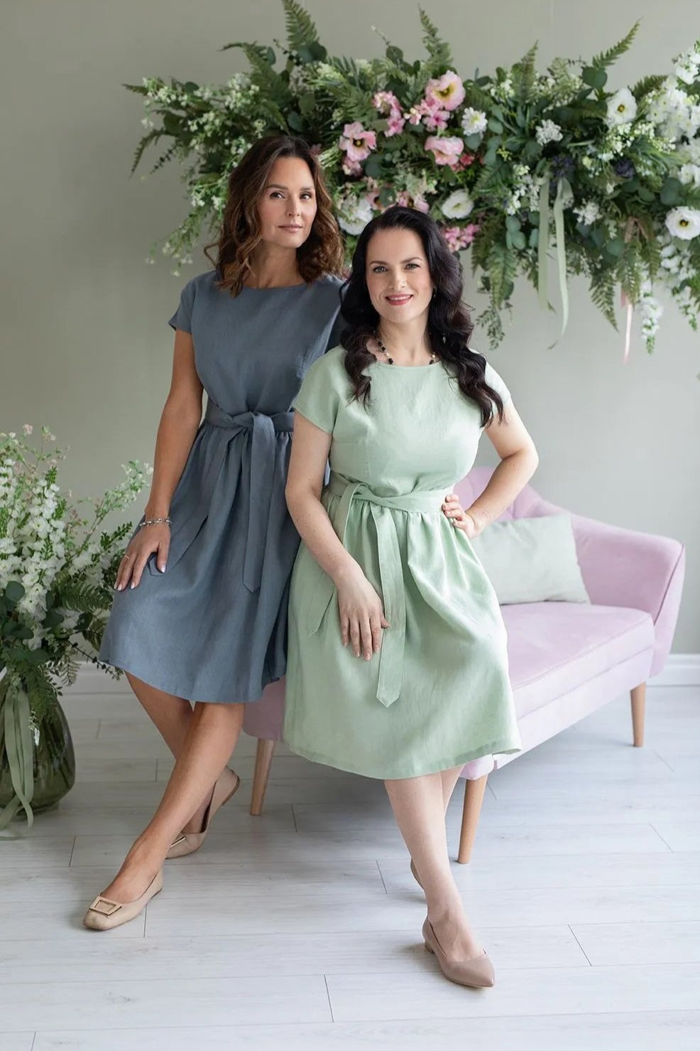 Two women in belted linen dresses, mint green and grey, posing in a floral setting