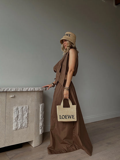 Woman in a brown sleeveless linen dress, straw hat, and straw tote bag, in a minimal room.