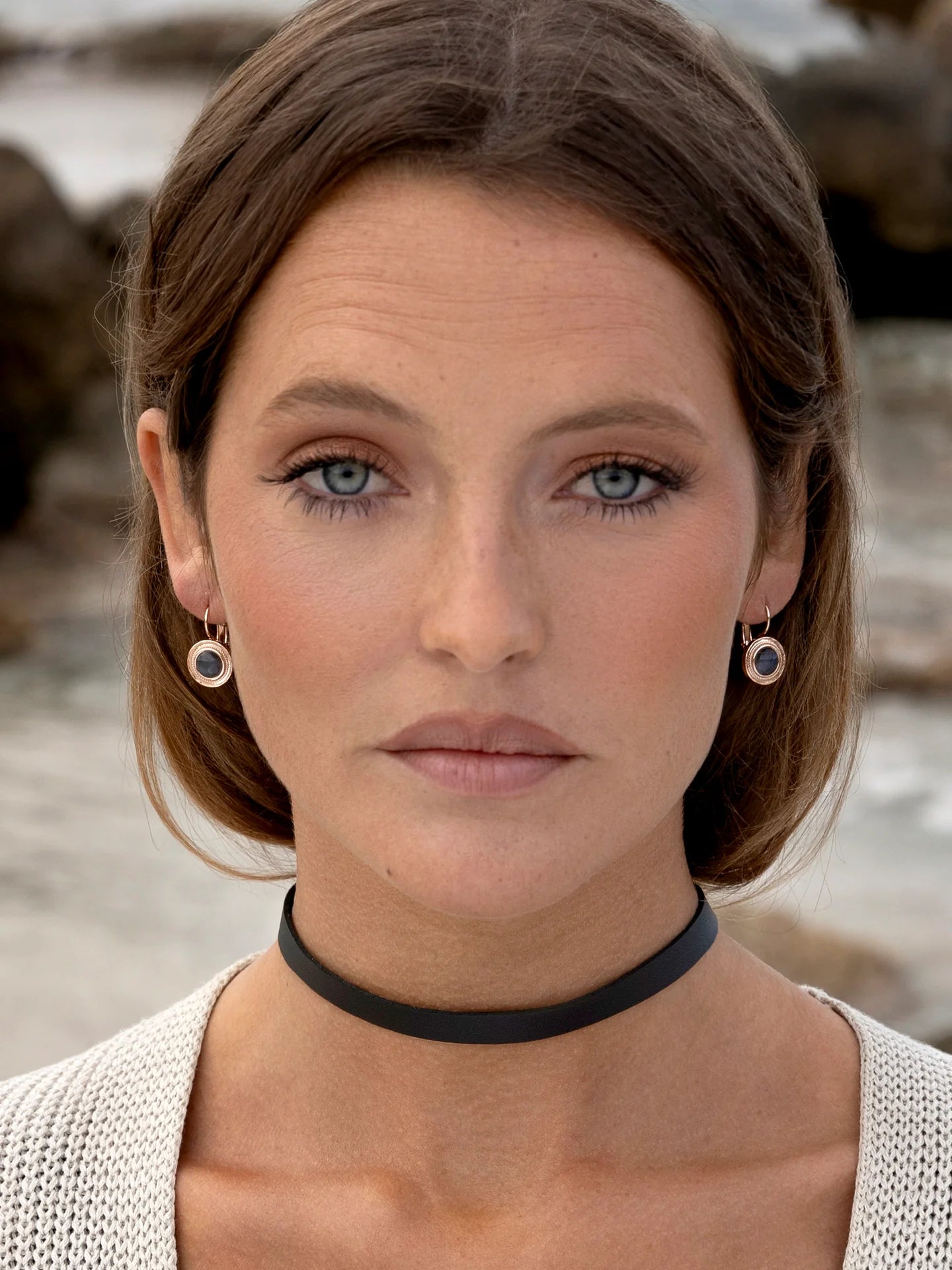 Woman wearing minimalist black choker, linen top, and drop earrings, outdoors by rocks