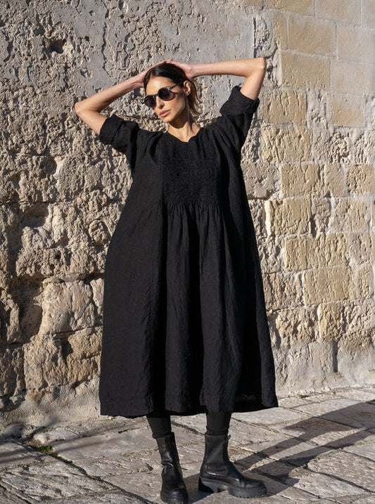 Woman wearing a loose black linen dress, sunglasses, and boots standing against a stone wall