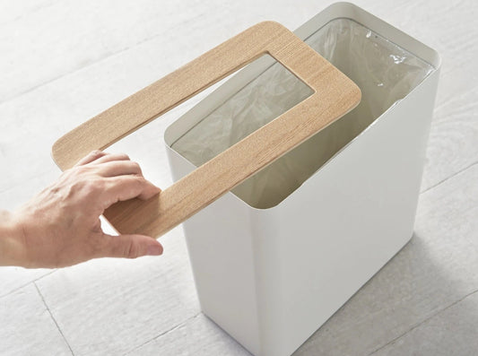 Modern white rectangular bin with wooden lid on a light floor, hand lifting lid