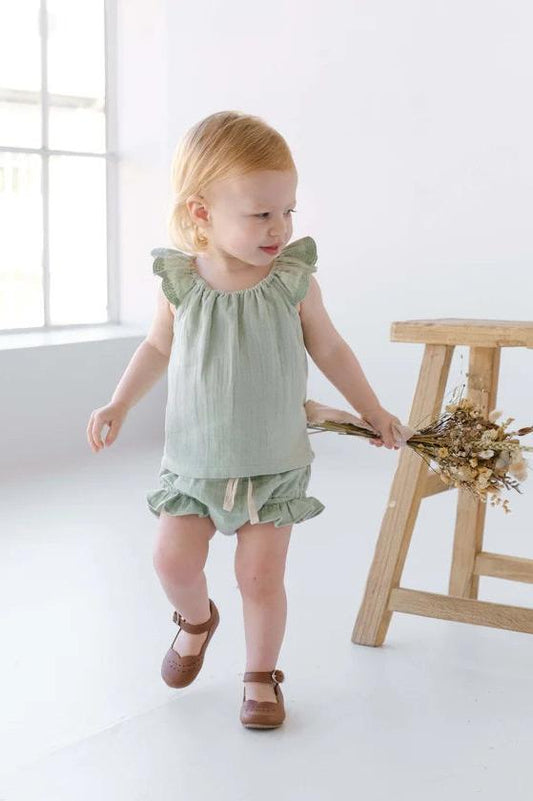 Toddler in sage green linen ruffle top and bloomers holding dried flowers indoors