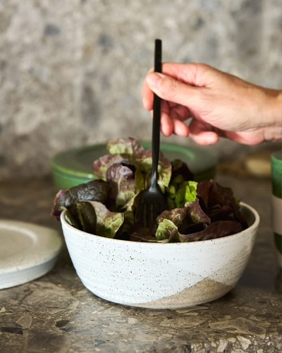 Hand holding black fork over fresh salad in ceramic bowl on stone counter