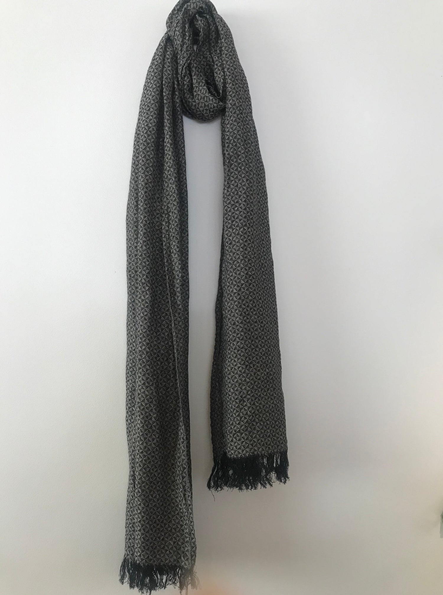 Grey patterned scarf with textured weave and fringed ends hanging against a white wall