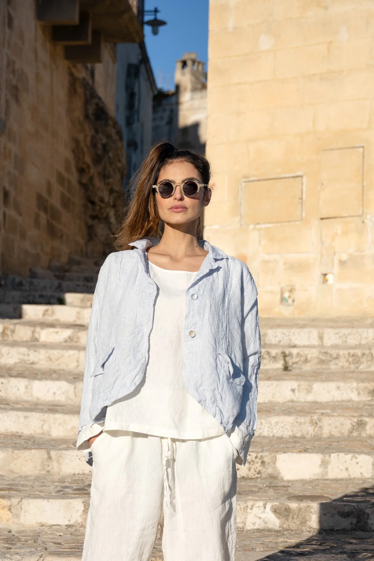 Woman wearing light blue linen jacket and white linen pants, standing outdoors in sunlight