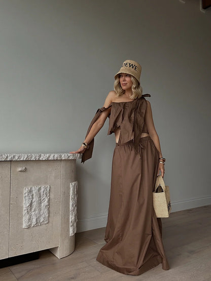 Woman wearing a brown linen maxi skirt and matching top, holding a straw bag indoors.