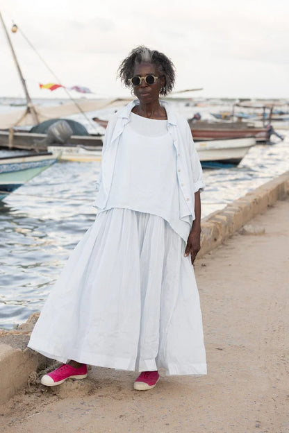 Woman in white linen shirt and skirt with pink shoes by waterfront, boats in background
