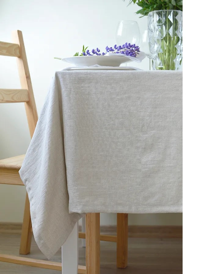 Natural linen tablecloth draped over dining table with plates and purple flowers