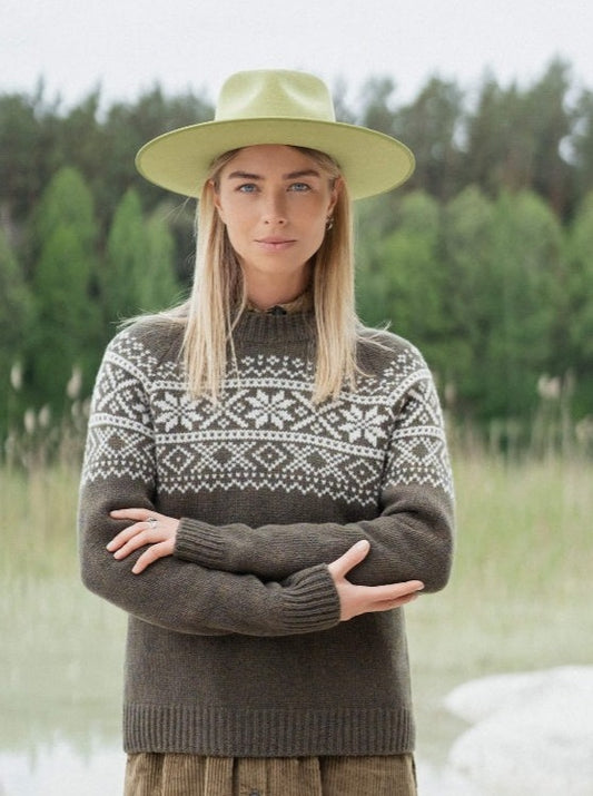 Woman in green hat and Nordic pattern sweater, standing outdoors by lake and trees
