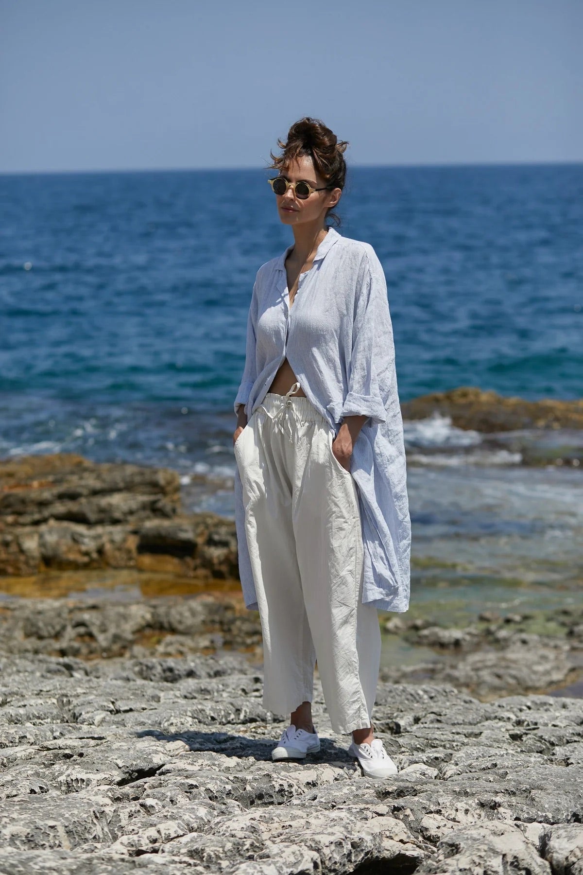 Woman in loose white linen shirt and pants standing on rocky shore with the ocean in background