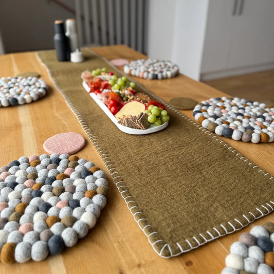 Olive felt table runner with white stitching on wooden dining table, surrounded by round felt placemats and a platter with cheese, crackers, grapes, and snacks.
