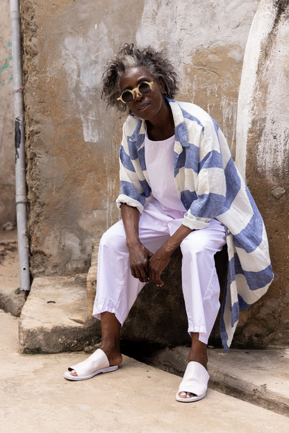 Woman in oversized blue and white linen shirt, white pants, and sandals sitting outdoors