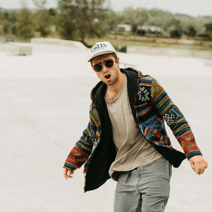 Man wearing round wooden sunglasses, colorful jacket, and cap outdoors
