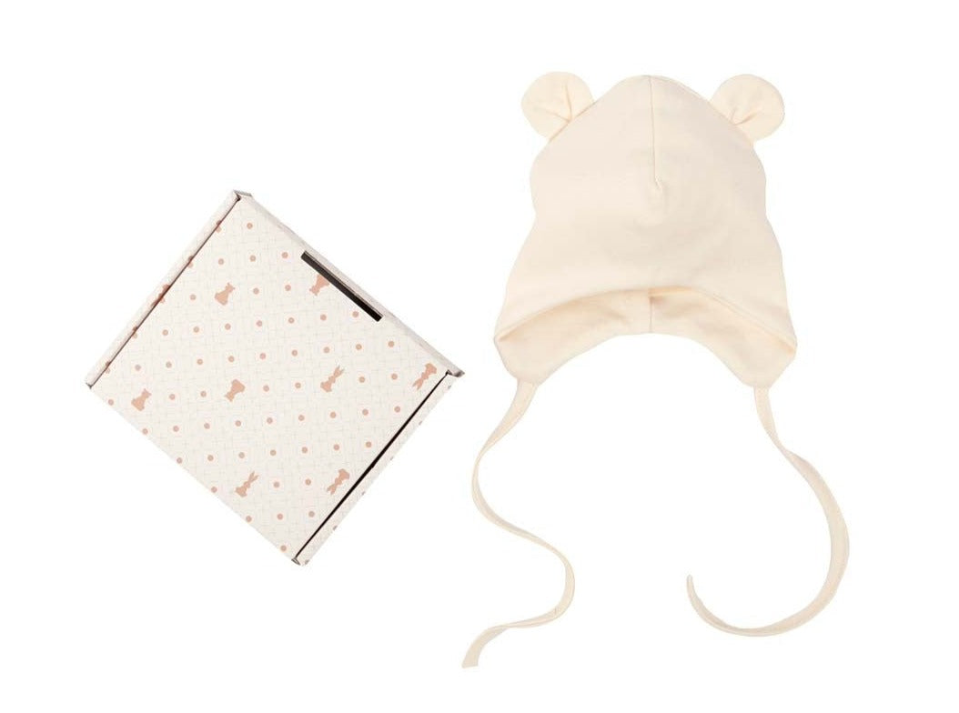 Beige baby hat with bear ears and ties, next to a patterned gift box on white background