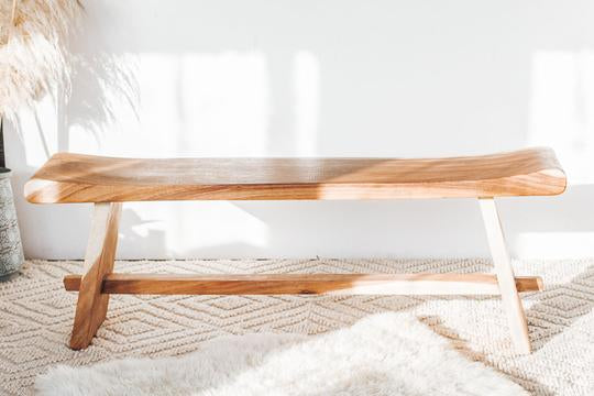 Wooden bench with a natural finish on a textured rug in a sunlit, minimalist room.